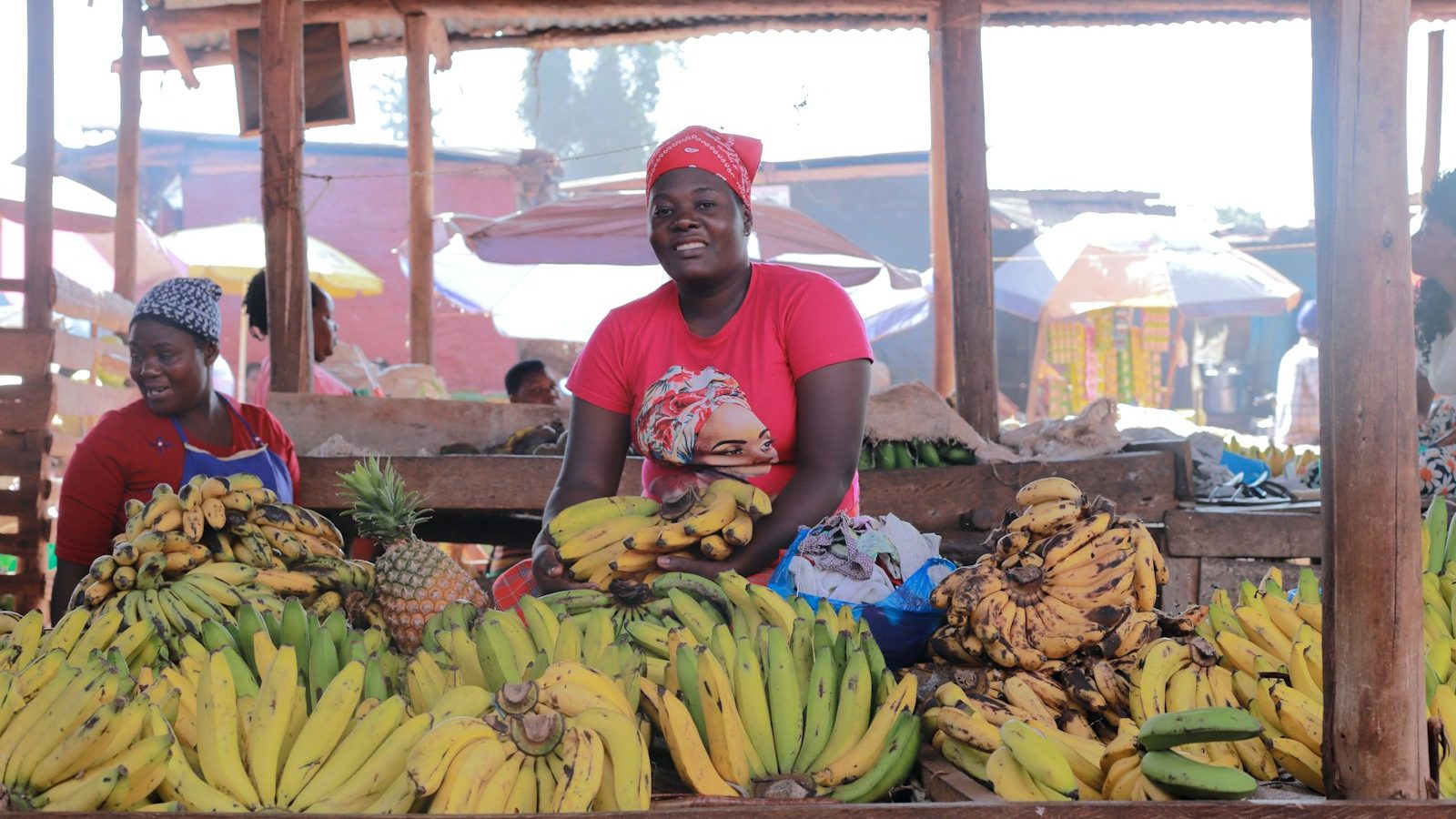 A trader standing behind a stall of bananas and pineapples in a Ugandan market